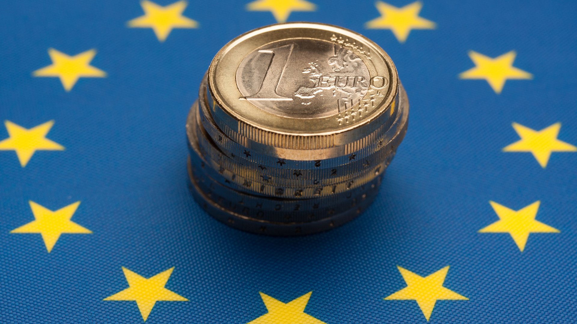 A pile of one Euro coins standing in the middle of the European Union flag. Blue background with a ring of yellow stars.