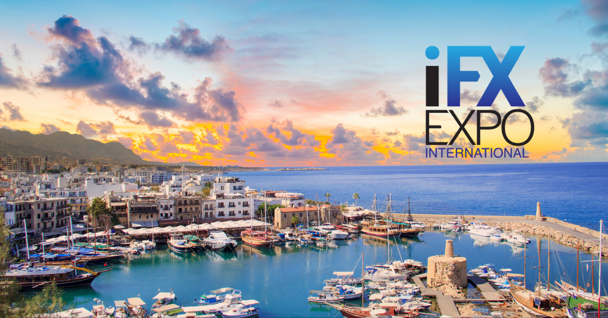 A vibrant harbor scene during sunset with a marina full of yachts and sailboats, surrounded by historic Mediterranean-style buildings. In the background, dramatic clouds reflect warm orange and purple hues. The sea stretches out to the horizon. On the right side of the image, the logo